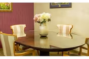 A dining area with a polished wooden oval table surrounded by four upholstered chairs with striped fabric. A vase with a bouquet of flowers is placed in the center of the table. The walls are painted in light beige and maroon colors, and a framed picture is partially visible on the left wall.