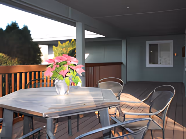 A covered outdoor patio area with a hexagonal table and four metal chairs. A vase with pink flowers is placed on the table. The patio has wooden flooring and railing, with trees visible in the background.