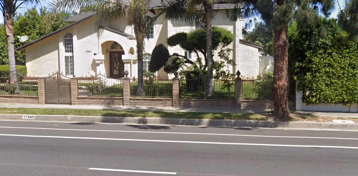 Front view of Devonshire Elderly Care facility with gate, fence, and trees along sidewalk.