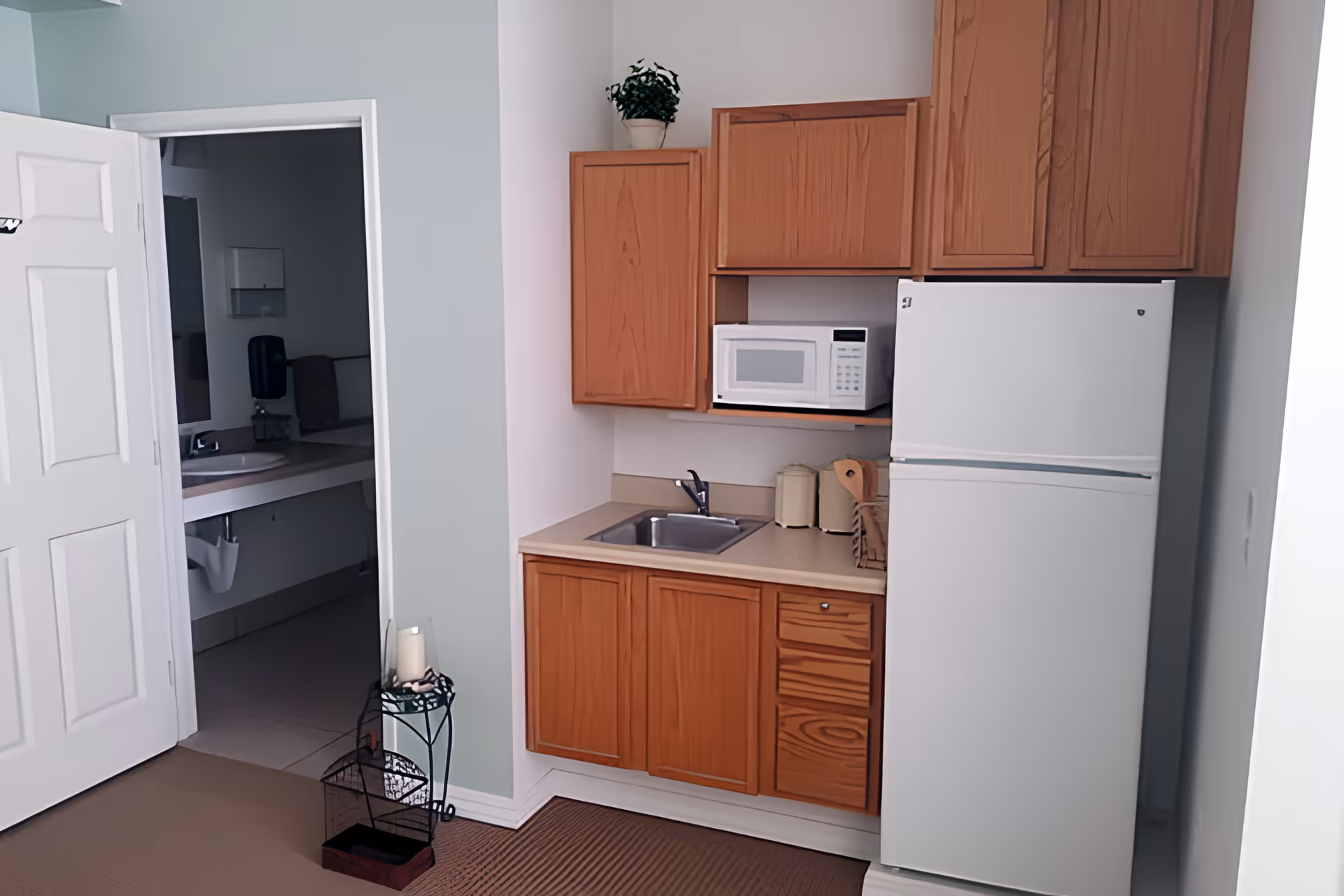 Small kitchenette area with wooden cabinets, a white microwave, a white refrigerator, and a sink. To the left, an open door reveals a bathroom with a sink and mirror. A small plant is placed on top of the upper cabinet, and a decorative candle holder with a candle is on the floor near the door.
