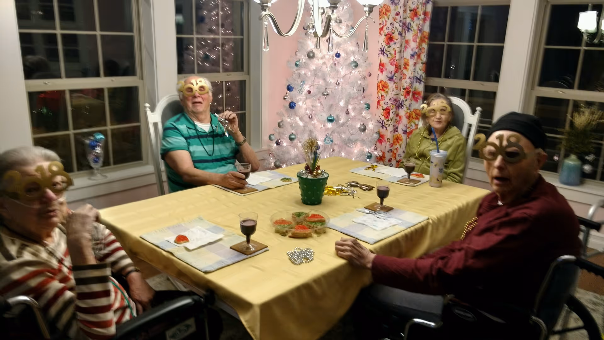 Four elderly people wearing '2018' novelty glasses sitting around a dining table with a yellow tablecloth. The table has placemats, small glasses with dark liquid, and some snacks. Behind them is a white Christmas tree with blue and silver ornaments, and floral curtains on the windows.