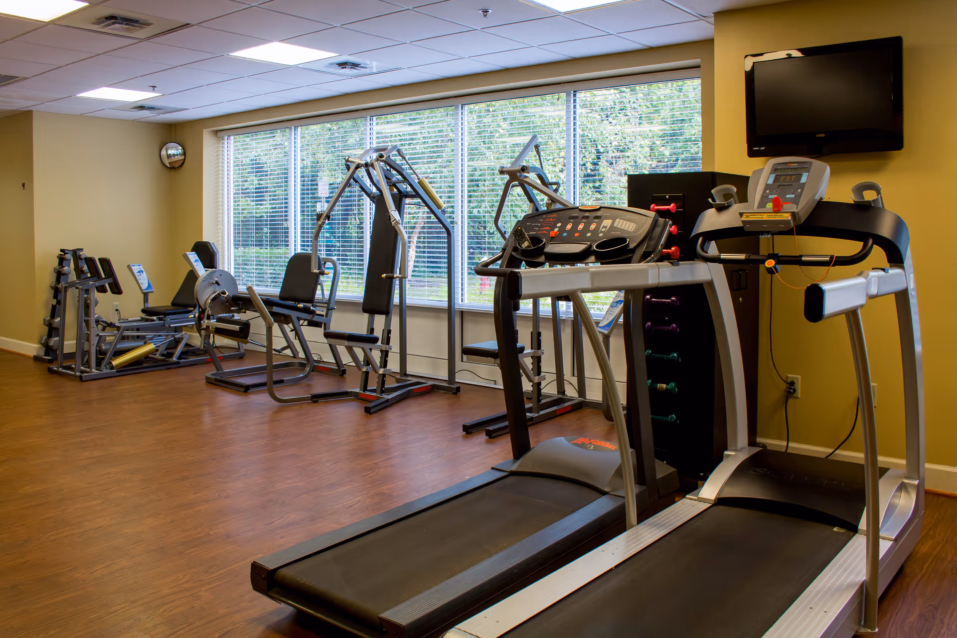 A fitness room with exercise equipment including two treadmills, weight machines, and a rack of dumbbells. Large windows with blinds allow natural light to enter the room, and a wall-mounted TV is visible above the treadmills. The floor is wood, and the walls are painted yellow.