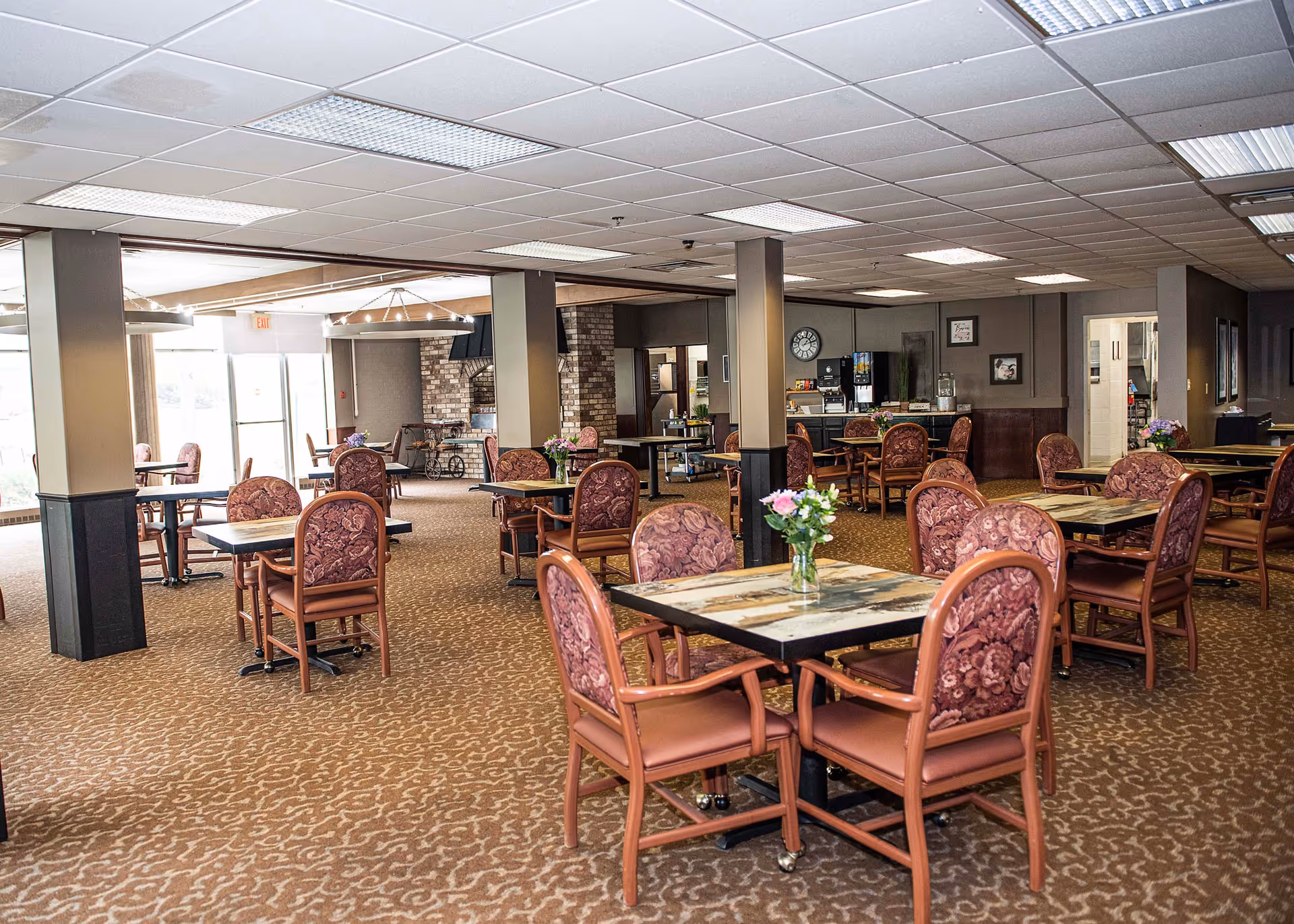 A spacious dining room with multiple tables and chairs arranged neatly. Each table has a small vase with flowers. The room has a carpeted floor, a drop ceiling with fluorescent lights, and large windows letting in natural light. In the background, there is a beverage station and a brick fireplace.