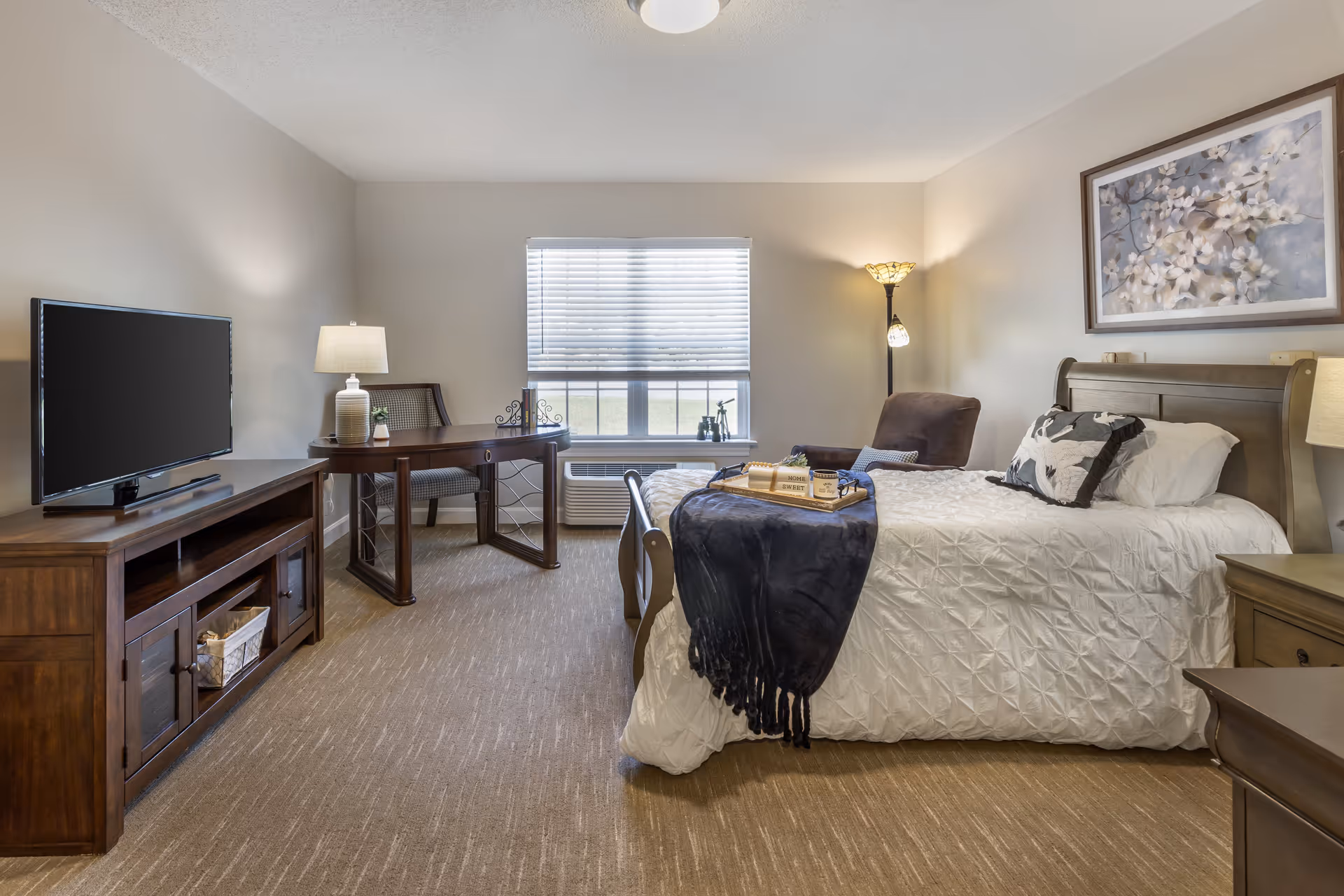 A cozy bedroom in a senior living facility featuring a bed with white bedding and decorative pillows, a dark blue throw blanket, a wooden nightstand with a lamp, a comfortable armchair, a wooden desk with a chair and lamp, a flat-screen TV on a wooden TV stand, and a window with blinds letting in natural light. A framed floral artwork hangs above the bed.