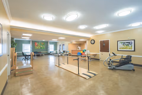 A spacious rehabilitation or therapy room with exercise equipment including parallel bars for walking practice, an elliptical machine, therapy balls, and a treatment table. The room has beige walls, bright ceiling lights, framed artwork, and windows letting in natural light.