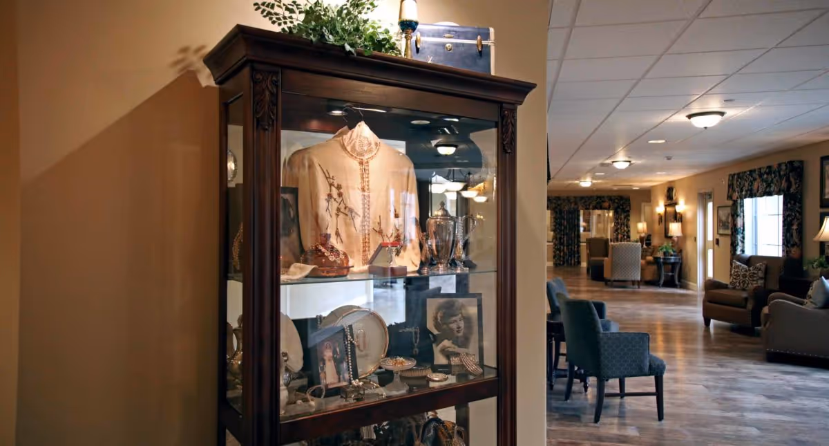 A wooden display cabinet filled with vintage items including a cream-colored embroidered jacket, framed black and white photographs, jewelry, and silverware. The cabinet is positioned in a hallway with wooden flooring and several upholstered chairs and sofas along the walls. The hallway is warmly lit with ceiling lights and wall sconces, and windows with floral curtains are visible.