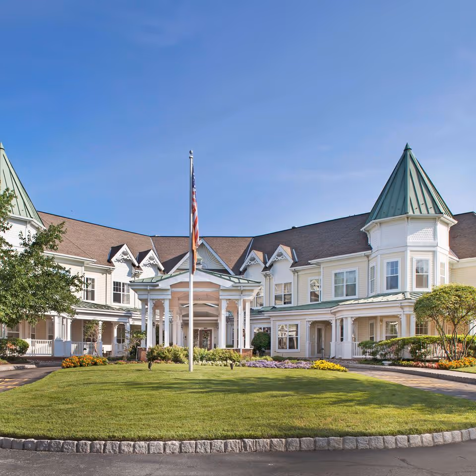 Front exterior view of a senior living facility with a circular driveway, a well-maintained lawn, flower beds, and an American flag on a flagpole in the center. The building has white siding, green roofs, and multiple windows under a clear blue sky.