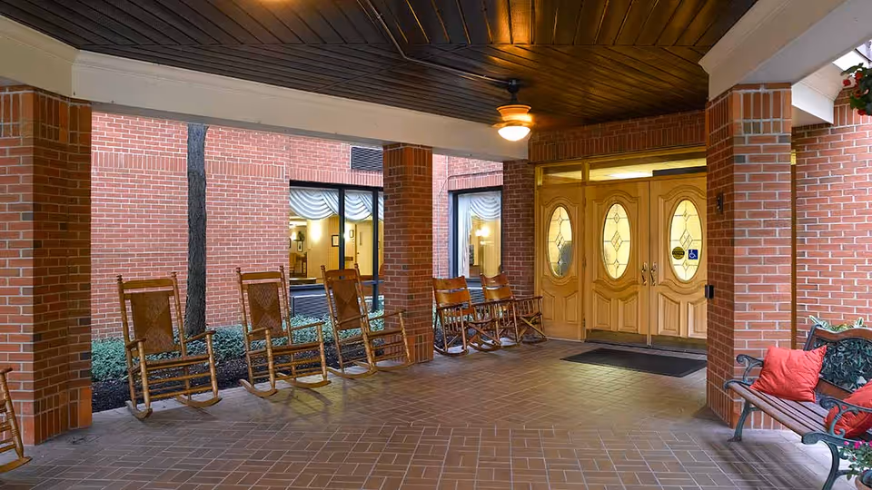 Covered entrance area of a senior living facility with brick pillars and walls, wooden double doors with oval glass panels, several wooden rocking chairs lined up along the wall, and a bench with red cushions on the right side.