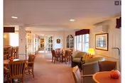 Bright assisted-living common area with dining tables, chairs, and lounge seating by windows.