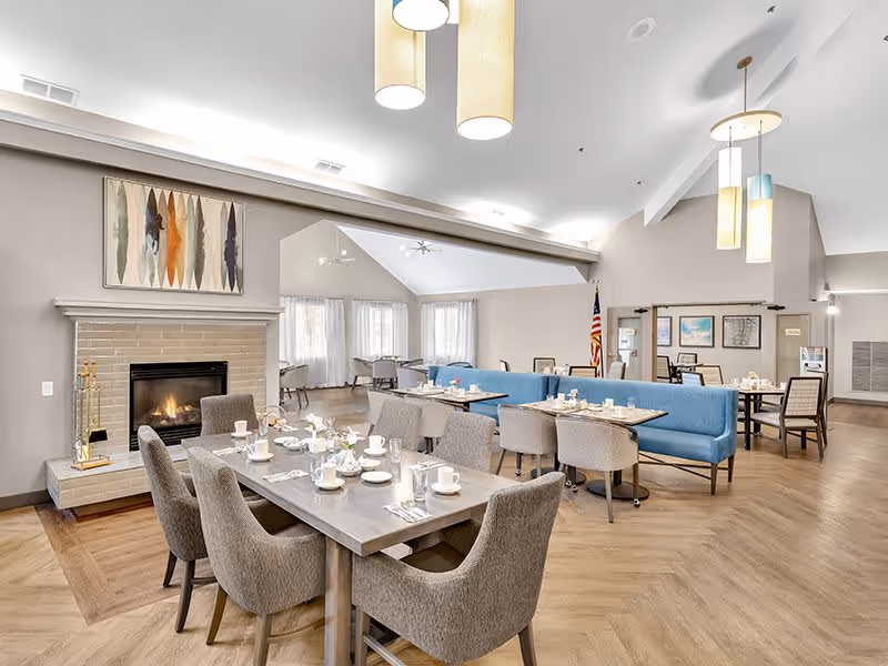 A spacious dining room with multiple tables set for a meal, featuring cups, plates, and utensils. The room has a modern design with light wood flooring, a fireplace on the left wall, and a blue cushioned bench seating area. Pendant lights hang from the ceiling, and there are large windows with sheer curtains allowing natural light to fill the space. An American flag is visible in the background near framed artwork on the walls.