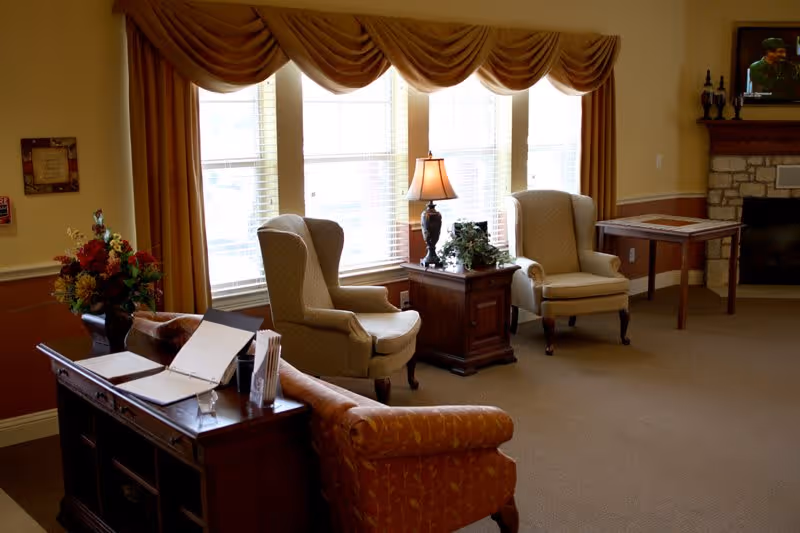 A cozy living room area with two beige upholstered armchairs, a wooden side table with a lamp and plant, a wooden console table with papers and a flower arrangement, large windows with brown drapes, and a wall-mounted TV above a stone fireplace.