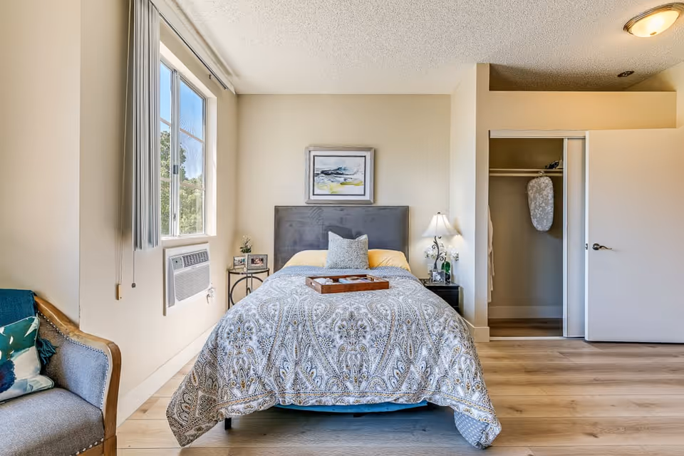 A bright bedroom with a bed centered against the wall, featuring a patterned bedspread and a decorative pillow. There is a framed abstract painting above the headboard. To the right of the bed is a nightstand with a lamp and some items. An open closet with sliding doors is visible on the right side of the room. On the left side, there is a window with vertical blinds and an air conditioning unit below it. A cushioned chair with a decorative pillow is partially visible in the left foreground. The room has light-colored walls and wood flooring.
