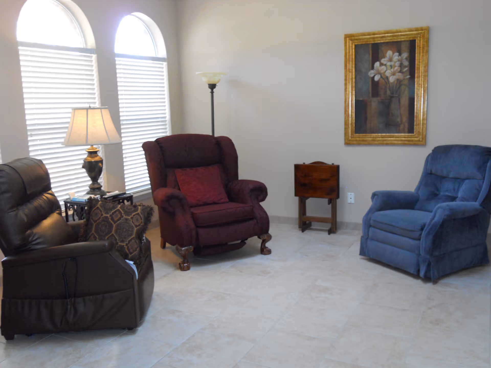 A cozy living room area with three recliner chairs in black, burgundy, and blue arranged in a semi-circle. There is a side table with a lamp and books next to the black chair, a floor lamp behind the burgundy chair, and a small wooden table against the wall. Two arched windows with white blinds let in natural light, and a framed floral painting hangs on the beige wall.