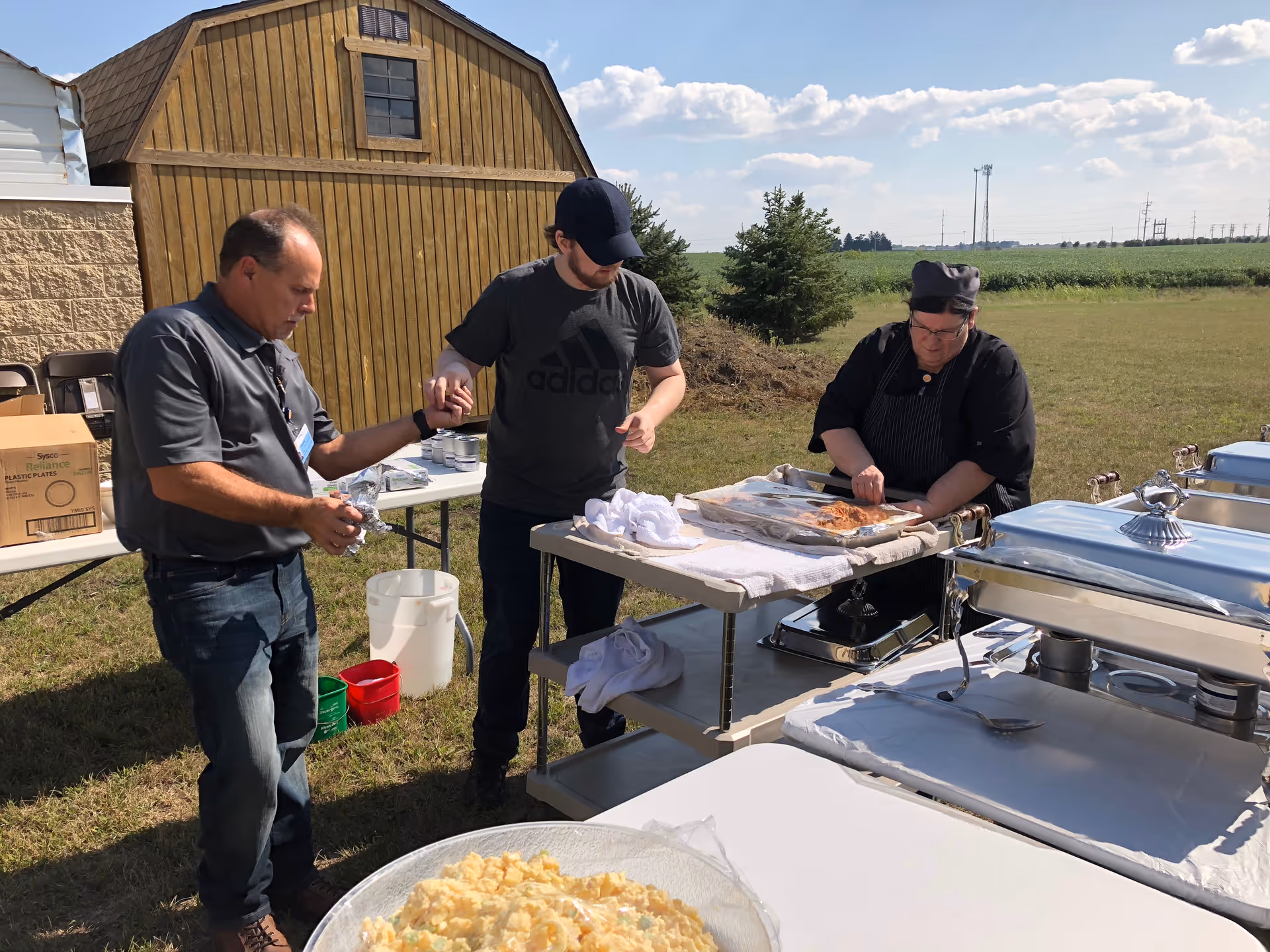 Three people outdoors near a wooden shed and tables set up with food and serving equipment. One person is wearing a chef's outfit and preparing food on a table, while the other two are standing nearby, one holding the other's hand. The setting is a grassy open area under a partly cloudy sky.