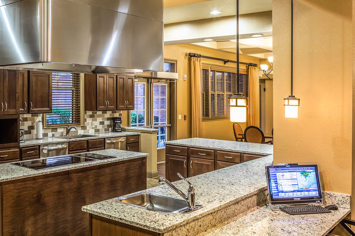 A modern kitchen area with granite countertops, a stainless steel sink, and a stovetop with a large stainless steel range hood. Wooden cabinets line the walls, and a coffee maker is visible on the counter. In the background, there is a dining area with a window covered by blinds and curtains, and a computer monitor with a keyboard is placed on the counter in the foreground.