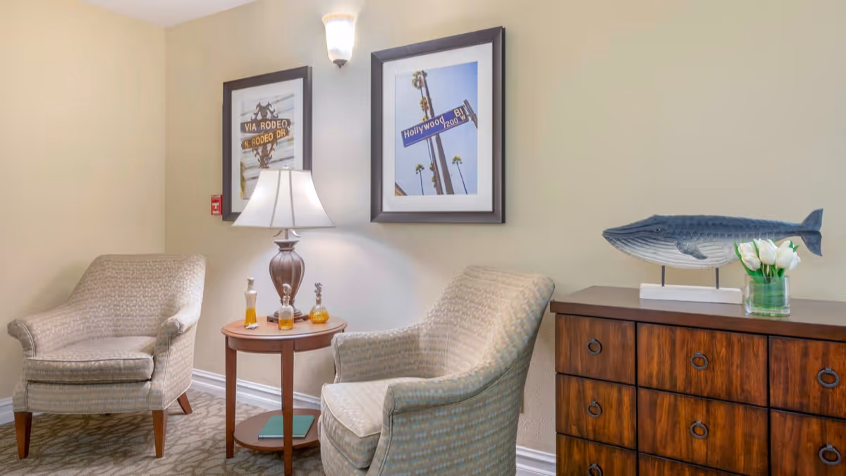 A cozy sitting area with two patterned armchairs facing each other, a round wooden side table with a lamp and decorative bottles, two framed street sign pictures on the wall, and a wooden dresser with a whale sculpture and a vase of white tulips.