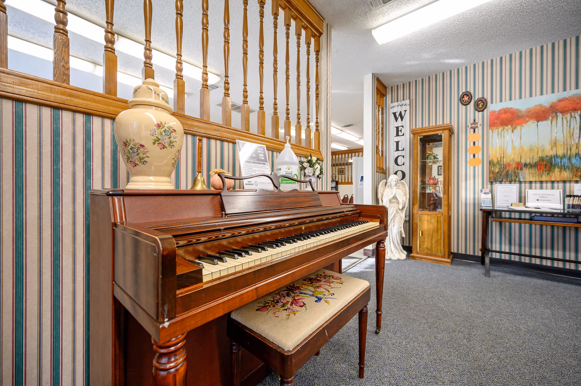 Interior view of a senior living facility featuring a wooden piano with a floral cushioned bench. On top of the piano are a decorative vase, a bell, a hand sanitizer bottle, and some informational signs. The room has striped wallpaper, a 'WELCOME' sign, a white angel statue, a glass display cabinet, and a table with brochures and a colorful painting of trees on the wall.