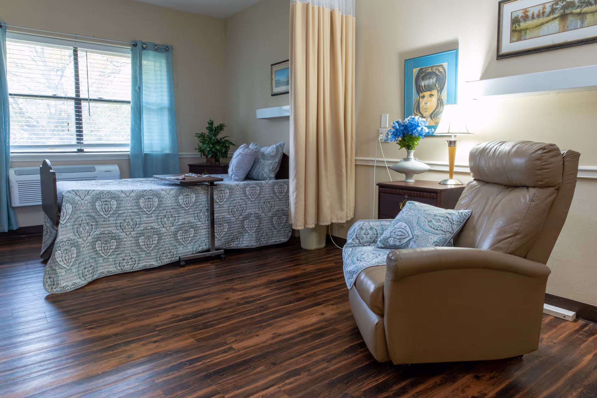 A bright retirement bedroom with a single bed, a tan recliner, bedside table and a window with blue curtains.