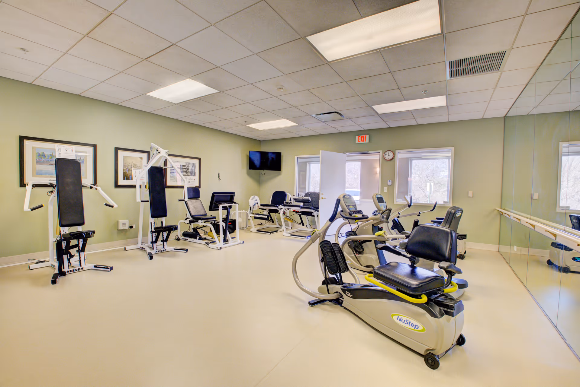 A spacious fitness room in a senior living facility with various exercise machines including NuStep recumbent cross trainers and strength training equipment. The room has light green walls, a large mirror on one side, framed pictures on the wall, a wall-mounted TV, and windows letting in natural light.