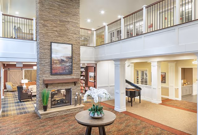 A spacious senior living facility common area with a tall stone fireplace in the center, a round table with a flower arrangement in front, a grand piano to the right, and seating areas with chairs and lamps on the left. The space has a second-floor balcony with railings overlooking the area.
