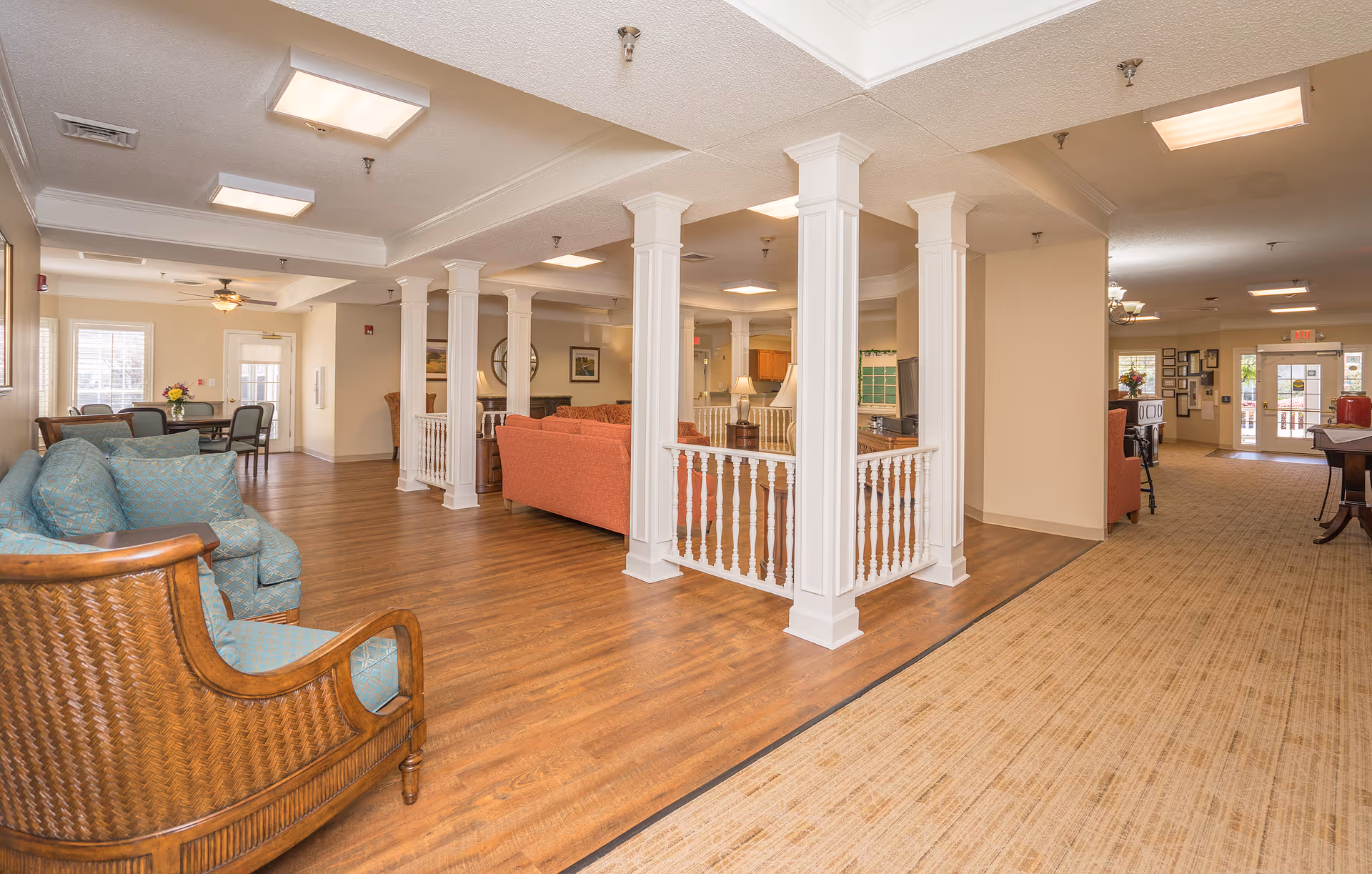 Open common lounge in a senior living facility with sofas, chairs, white columns, and a hallway leading to the entrance.