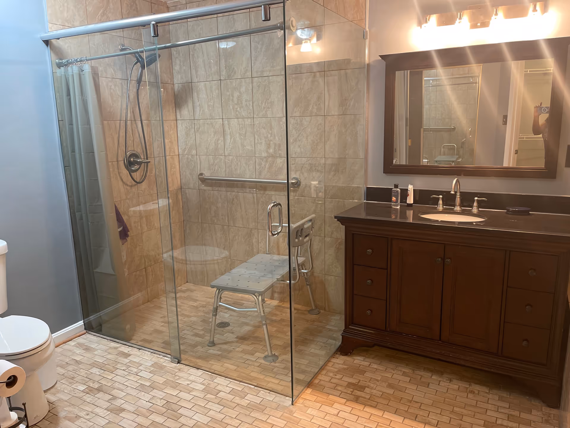 A bathroom with a glass-enclosed shower area featuring a shower chair and grab bar. The floor and shower walls are tiled in beige tones. There is a dark wood vanity with a black countertop, a sink, and a large mirror above it. A toilet with a roll of toilet paper is visible on the left side.