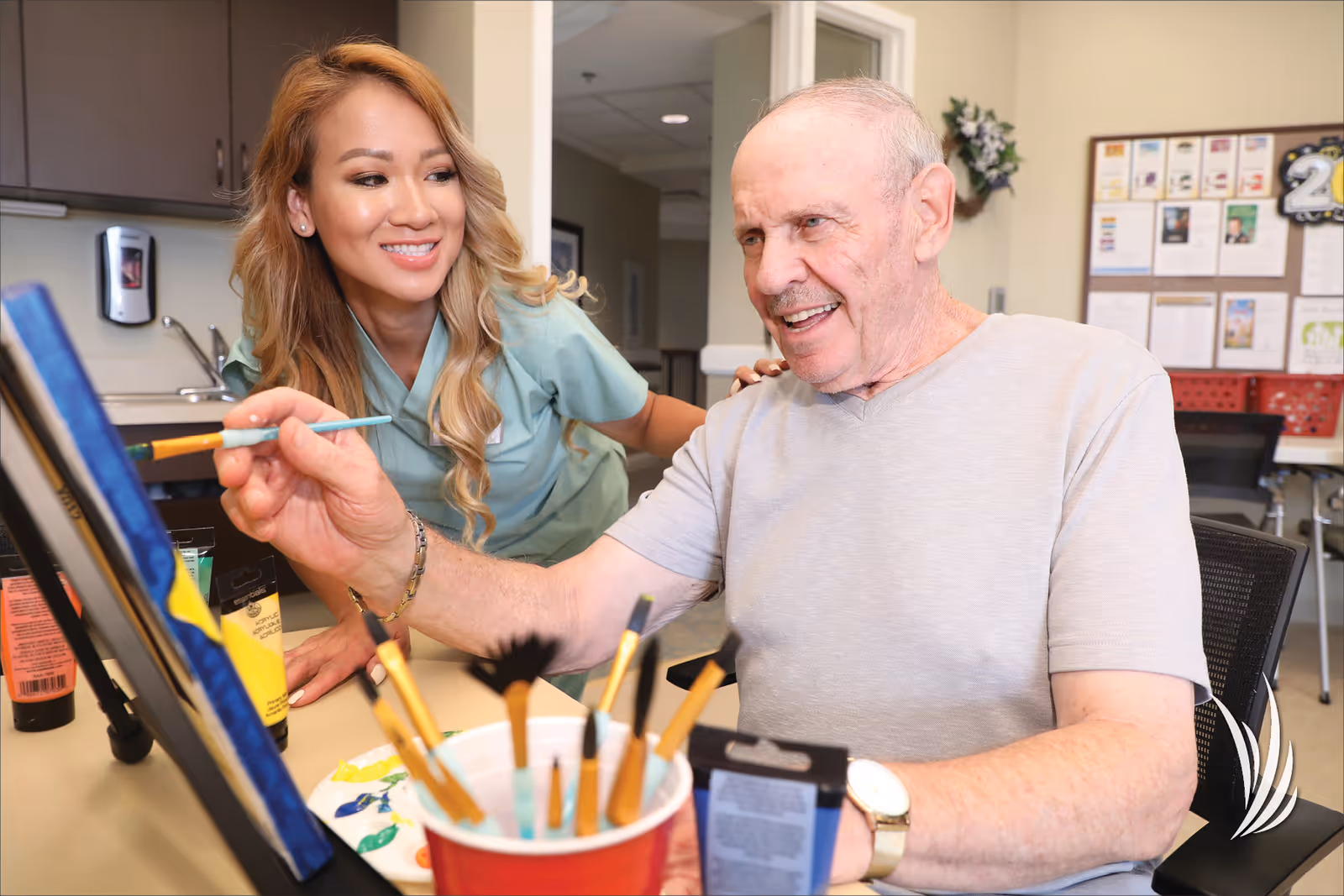 An elderly man painting on a canvas with a paintbrush while a smiling caregiver in scrubs watches and supports him in a well-lit room with art supplies on the table.