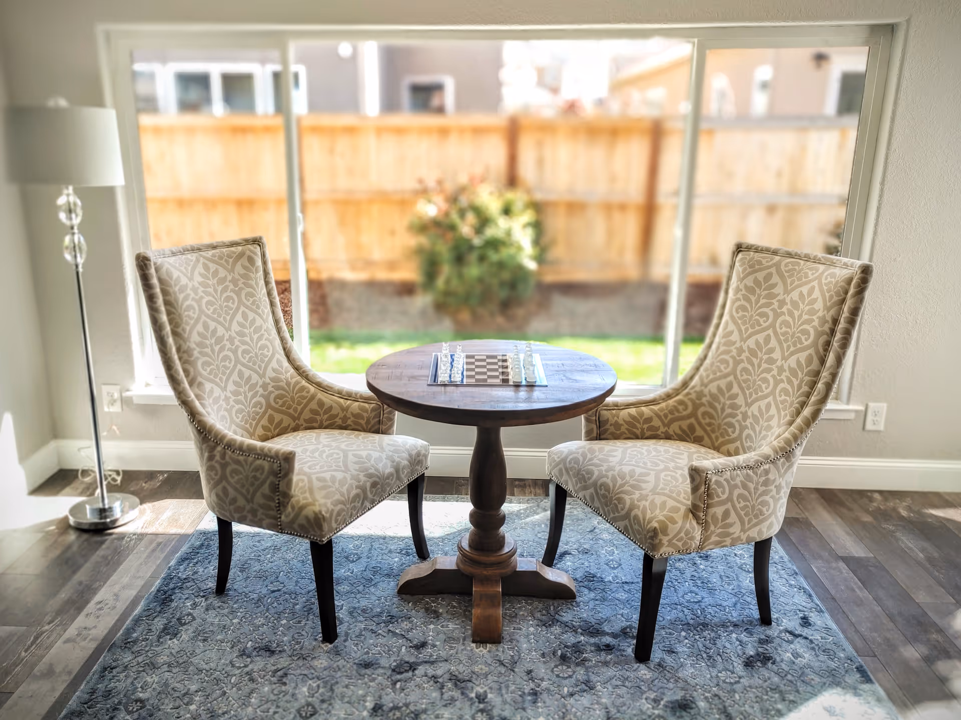 Two patterned upholstered chairs facing each other with a small round wooden table between them, set with a chessboard. The setting is in a bright room with large windows showing a fenced backyard with greenery outside. A tall floor lamp is positioned to the left of the chairs.