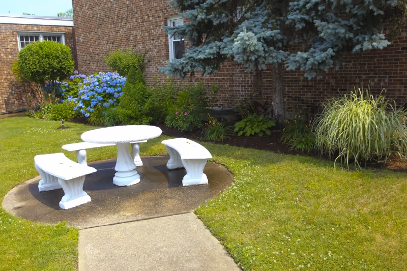 Outdoor garden area with a white round stone table and three matching curved benches on a concrete pad, surrounded by green grass, shrubs, and a brick building wall with windows in the background.