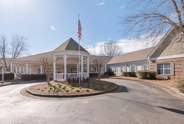 Exterior view of a single-story assisted living facility building with a circular driveway and a central gazebo-like entrance. An American flag is flying on a flagpole in the middle of the circular driveway. The building has a combination of brick and siding with multiple windows and some landscaping with small trees and bushes.