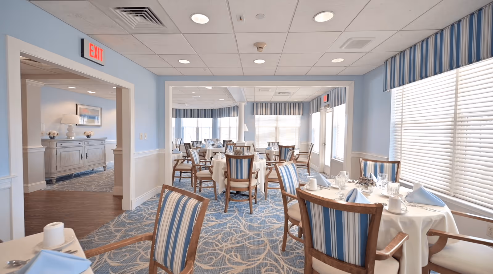 A bright dining room with round tables covered in white tablecloths, set with blue napkins, cups, and glassware. The chairs have wooden frames with blue and white striped upholstery. Large windows with blinds and matching striped valances let in natural light. The walls are painted light blue with white wainscoting, and the carpet has a blue and beige pattern. An exit sign is visible above a doorway leading to another room.