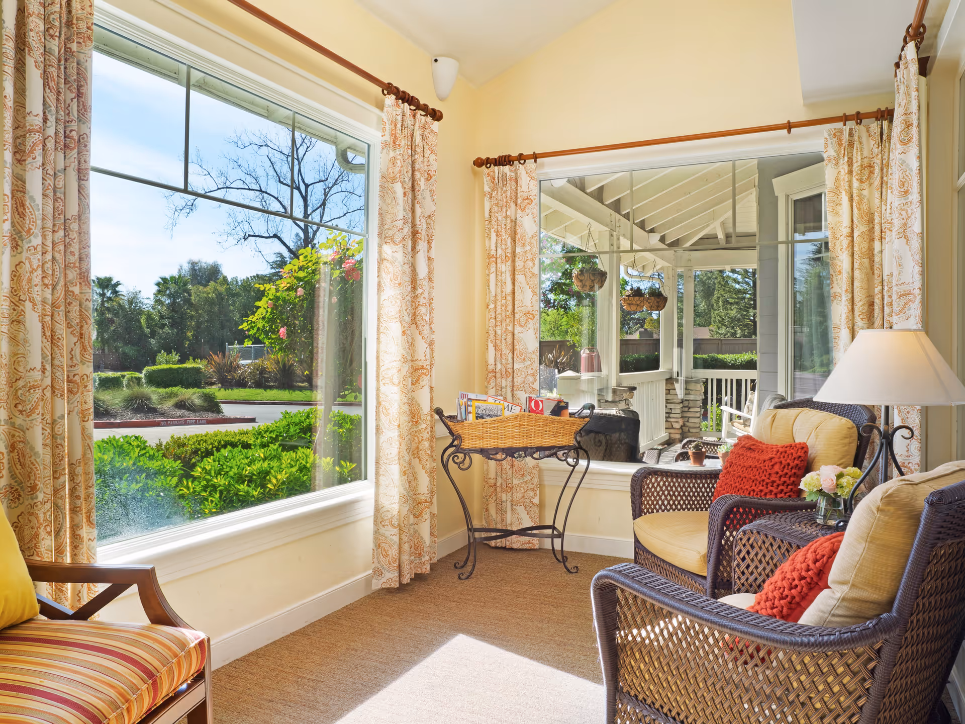 A bright and cozy sitting area with large windows showing a green outdoor garden. The room features wicker chairs with yellow cushions and red throw pillows, a small side table with a lamp and flowers, patterned curtains, and a basket stand holding magazines.