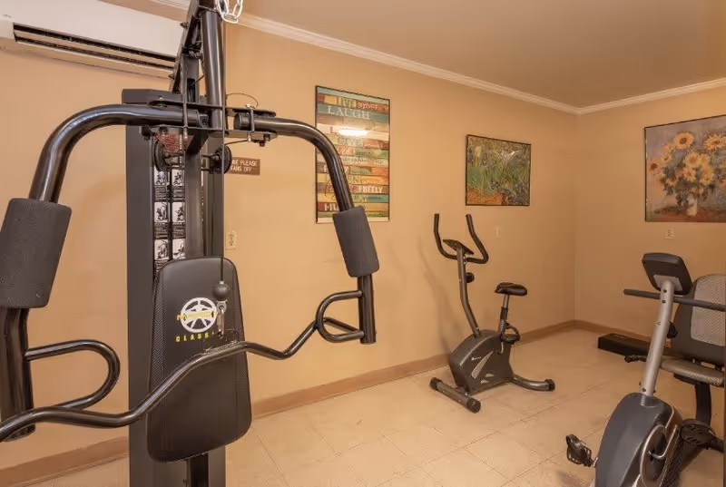 A small exercise room with fitness equipment including a multi-function weight machine, a stationary exercise bike, and a treadmill. The walls are beige with three framed pictures hanging, including a colorful motivational sign and two floral paintings.