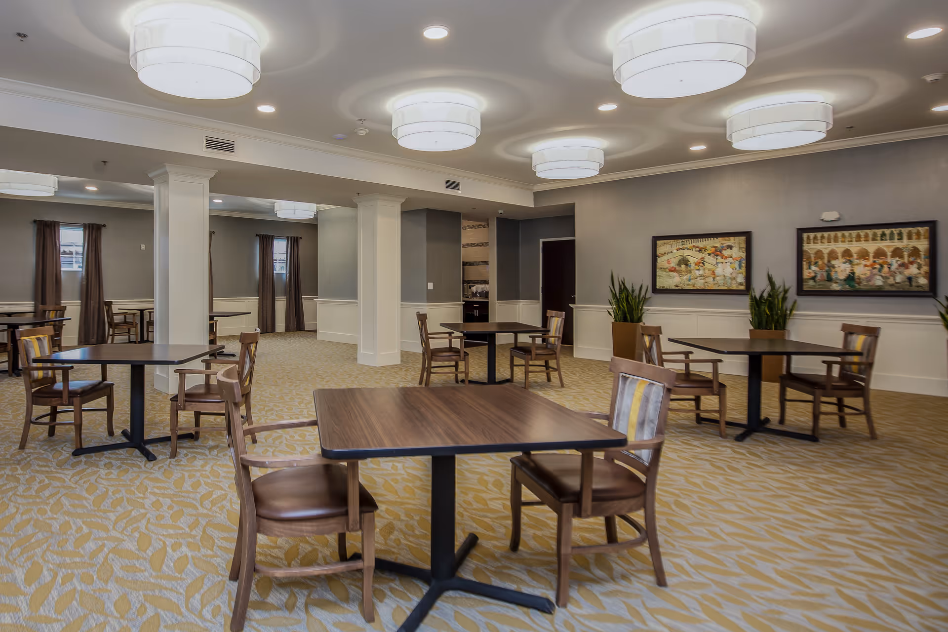 Spacious senior living dining room with multiple wood tables and chairs, decorative ceiling lights, plants, and framed artwork on the walls.