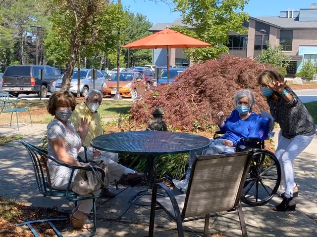 Four masked seniors—three seated around an outdoor metal table (one in a wheelchair) and one standing—waving in a sunny courtyard outside the care facility.