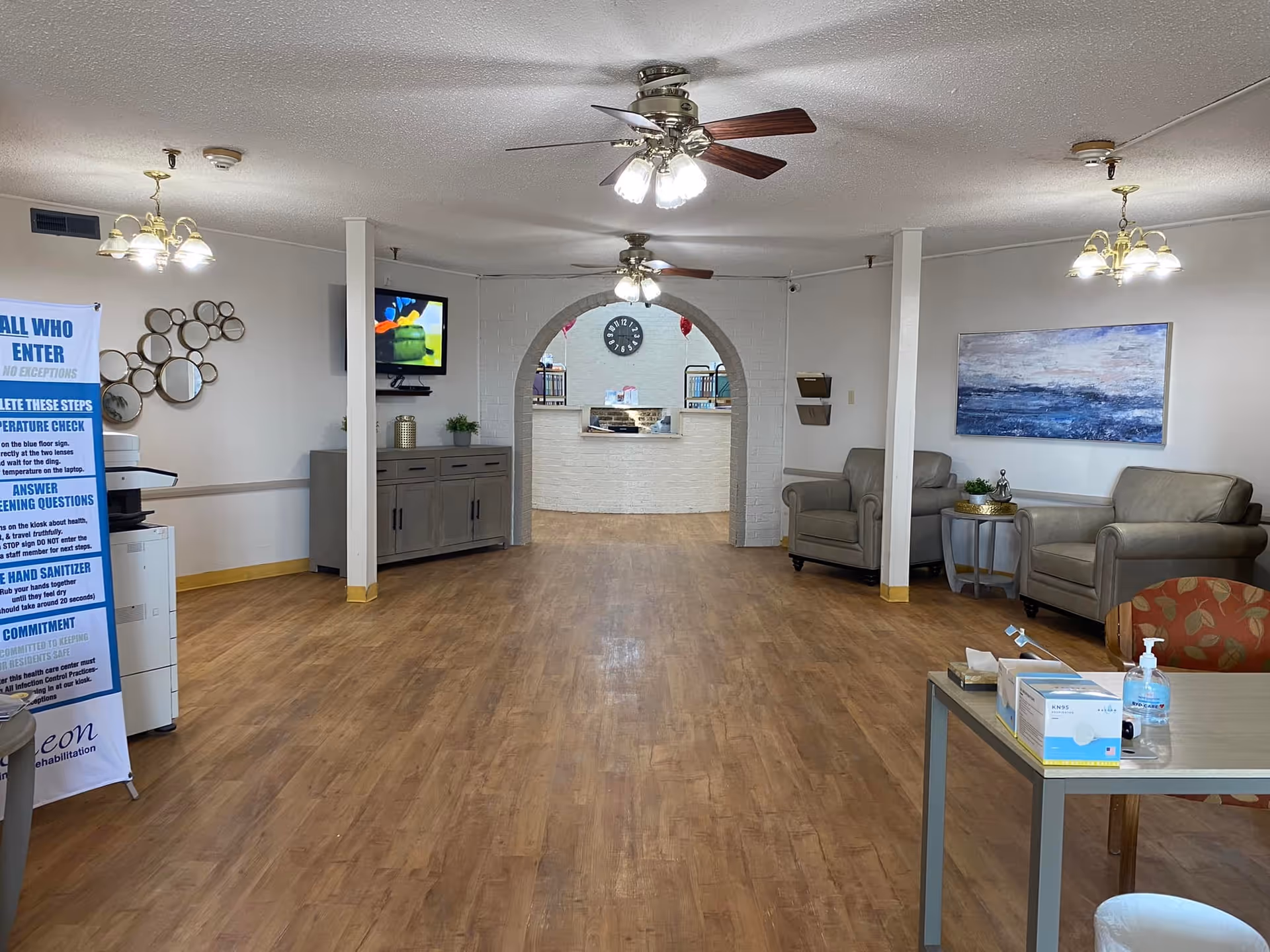 A spacious nursing and rehab facility common area with wooden flooring, two ceiling fans with lights, and comfortable seating including armchairs and a small table. There is a large archway leading to a reception desk with a clock on the wall behind it. A TV is mounted on the left wall above a cabinet, and a large blue and white informational sign is visible on the left side. The walls are decorated with a circular mirror arrangement and a large blue abstract painting. A table in the foreground holds hand sanitizer and boxes of face masks.