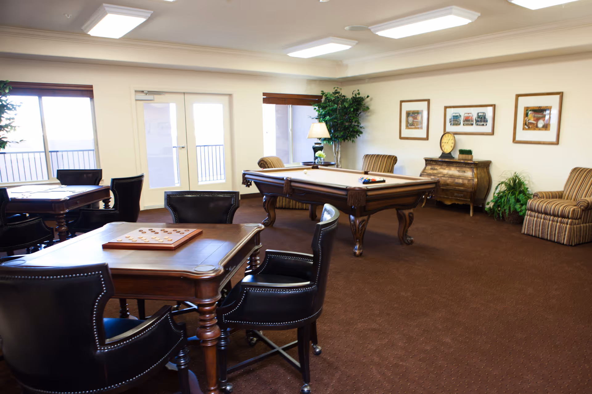 A spacious game room with a pool table in the center, surrounded by comfortable chairs and tables with board games. The room has large windows and glass doors letting in natural light, with framed pictures and plants decorating the walls and corners.