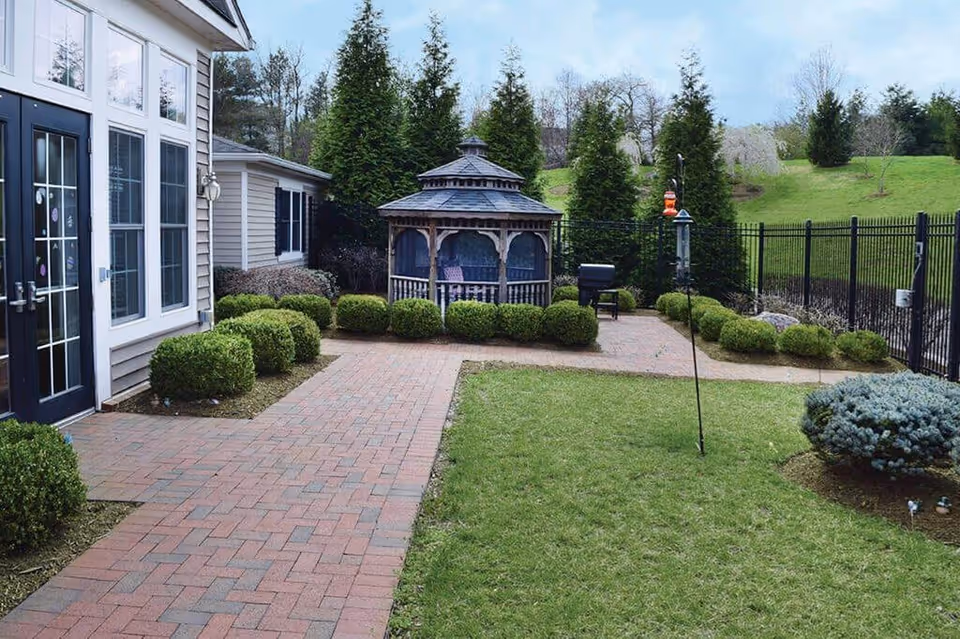 Outdoor view of a senior living facility courtyard with a brick pathway, manicured bushes, a grassy area, and a wooden gazebo. The area is enclosed by a black metal fence, and there are tall evergreen trees and a green hill in the background.