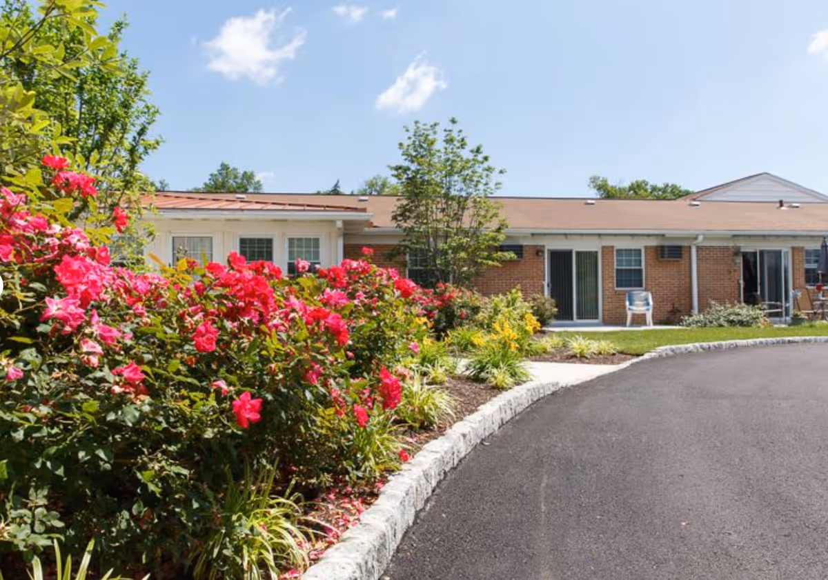 Exterior view of a single-story brick building with a curved paved driveway in front. There are vibrant pink flowers and green shrubs along the edge of the driveway, with a clear blue sky overhead.