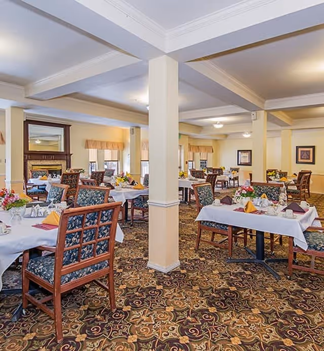 A spacious dining room with multiple tables covered in white tablecloths, each set with folded napkins, cups, and floral centerpieces. The room features patterned carpet, wooden chairs with floral upholstery, cream-colored walls, and several windows with beige valances. There is a fireplace with a mirror above it on one side of the room.