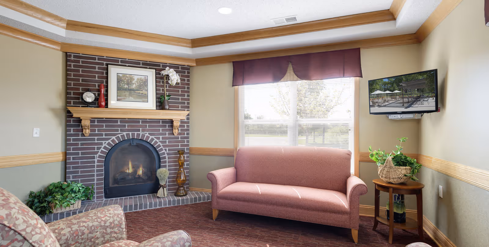 A cozy living room area with a brick fireplace, a pink upholstered sofa, an armchair with floral pattern, a small wooden side table with a basket of green plants, and a wall-mounted TV showing an outdoor patio scene. The room has beige walls with wooden trim and a large window with blinds and a purple valance.