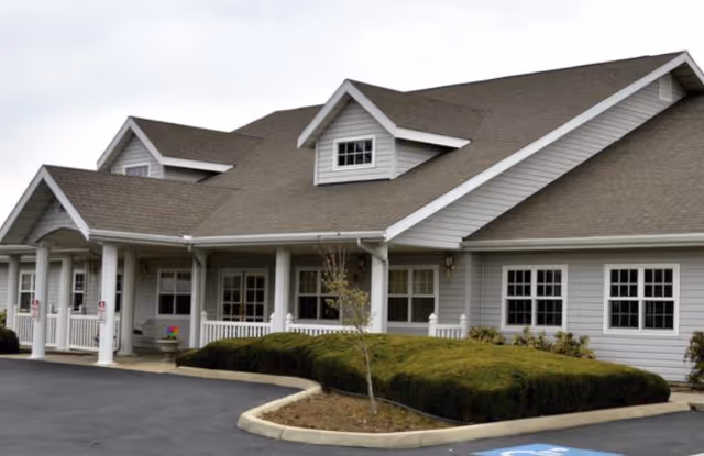Exterior view of a single-story building with gray siding and a brown shingled roof, featuring white columns and multiple windows. There is a small landscaped area with bushes and a young tree in front, and a paved parking lot with a handicap parking space visible.
