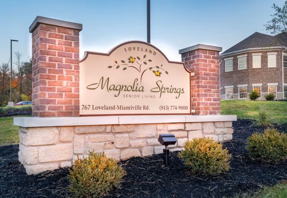 A large outdoor sign for Magnolia Springs Senior Living in Loveland, featuring the address 767 Loveland-Miamiville Rd. and a contact phone number. The sign is mounted on a stone and brick base with landscaping around it, and a building is visible in the background.