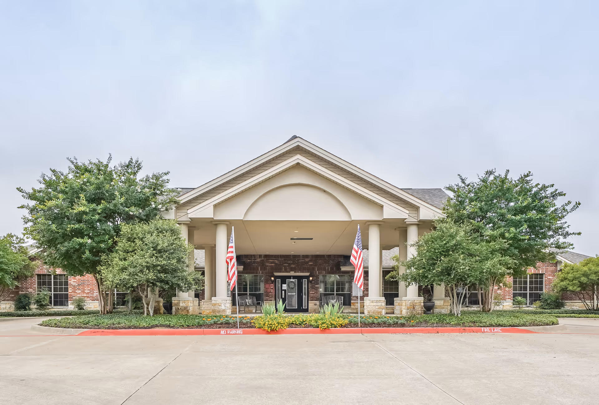 Front exterior view of TruCare Living Centers Palestine building with a covered entrance supported by columns, two American flags on either side, surrounded by trees and landscaping.