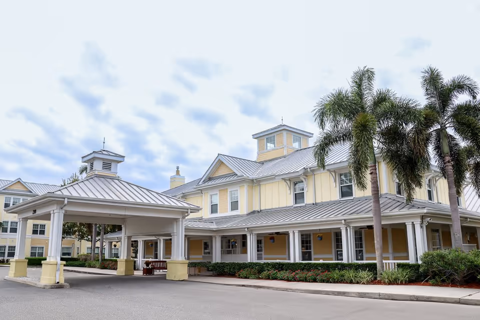 Exterior view of The Brennity at Melbourne Senior Living facility showing a large yellow and white building with a covered entrance, palm trees, and landscaped greenery under a cloudy sky.