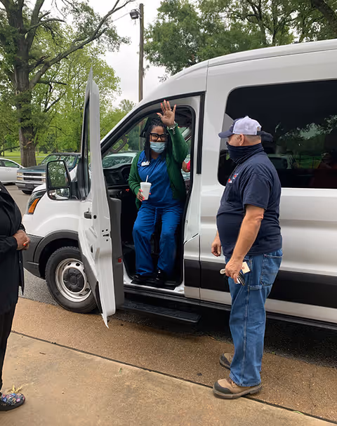 A woman wearing blue scrubs, a green cardigan, glasses, and a face mask is sitting in the driver's seat of a white van with the door open, waving. A man wearing a navy blue shirt, jeans, a cap, and a face covering stands outside the van holding a phone. Another person is partially visible on the left side of the image. The scene takes place outdoors with trees and parked cars in the background.