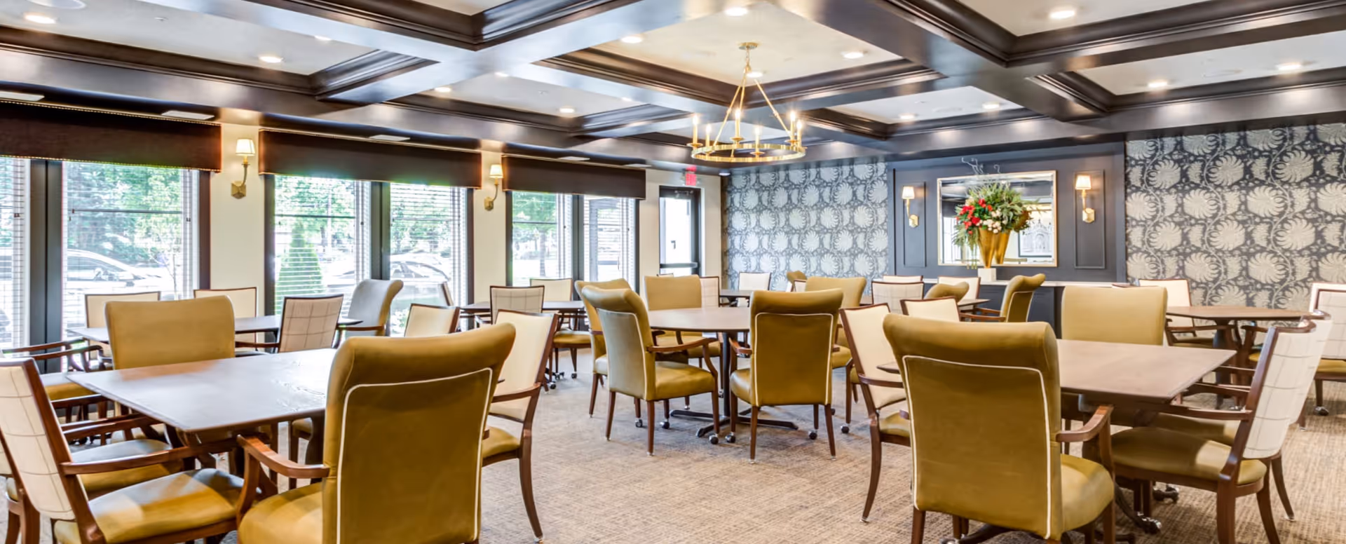A spacious dining room in a senior living facility with multiple wooden tables surrounded by cushioned chairs. Large windows with blinds allow natural light to fill the room. The walls feature patterned wallpaper and sconces with warm lighting. A decorative chandelier hangs from the ceiling, and a large floral arrangement is placed on a side table against the wall.