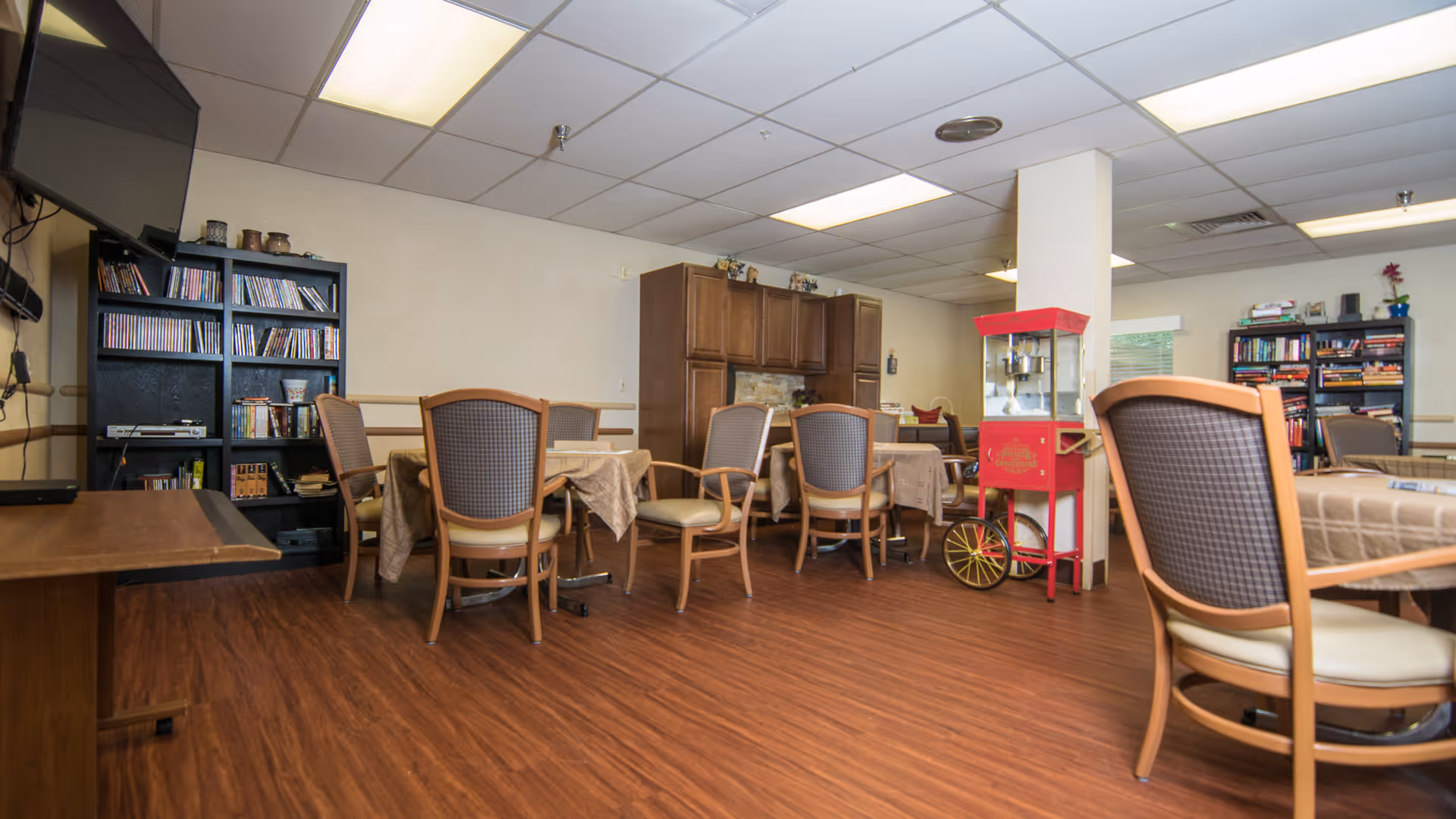 A common area in a senior living facility with multiple tables covered with beige tablecloths and surrounded by wooden chairs with cushioned seats and backs. There is a red popcorn machine near a white column in the center of the room. Bookshelves filled with books and DVDs line the walls. A large flat-screen TV is mounted on the left wall. The floor is wood laminate, and the ceiling has recessed lighting panels.