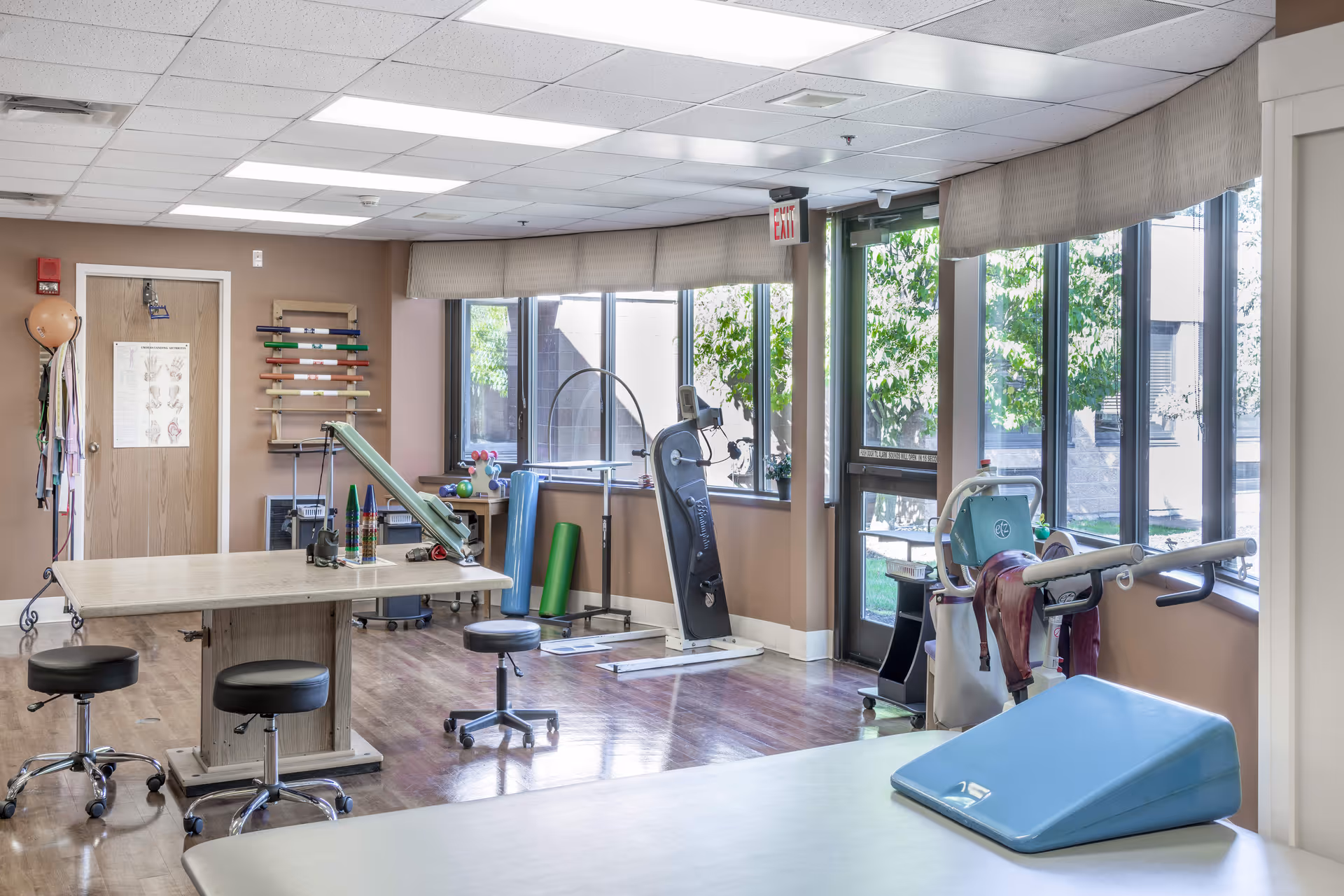 A bright therapy or rehabilitation room with large windows letting in natural light. The room contains various physical therapy equipment including exercise mats, stools on wheels, a therapy table, resistance bands, and other rehabilitation tools. The walls are beige, and the floor is wooden. There is an exit door with a red 'EXIT' sign above it.