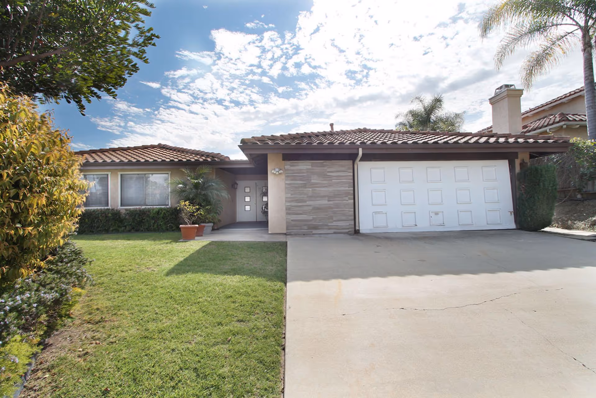 Front exterior of a single-story house with a two-car garage, concrete driveway, manicured lawn, potted plants, and a tiled roof under a partly cloudy sky.